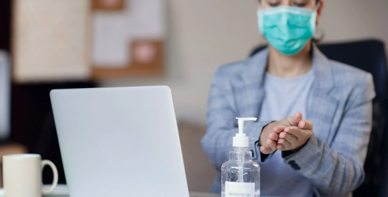 Woman wearing face mask while working in office with laptop and using hand alcohol gel or sanitizer bottle dispenser against corona virus Covid-19 / iStock photo Space_Cat