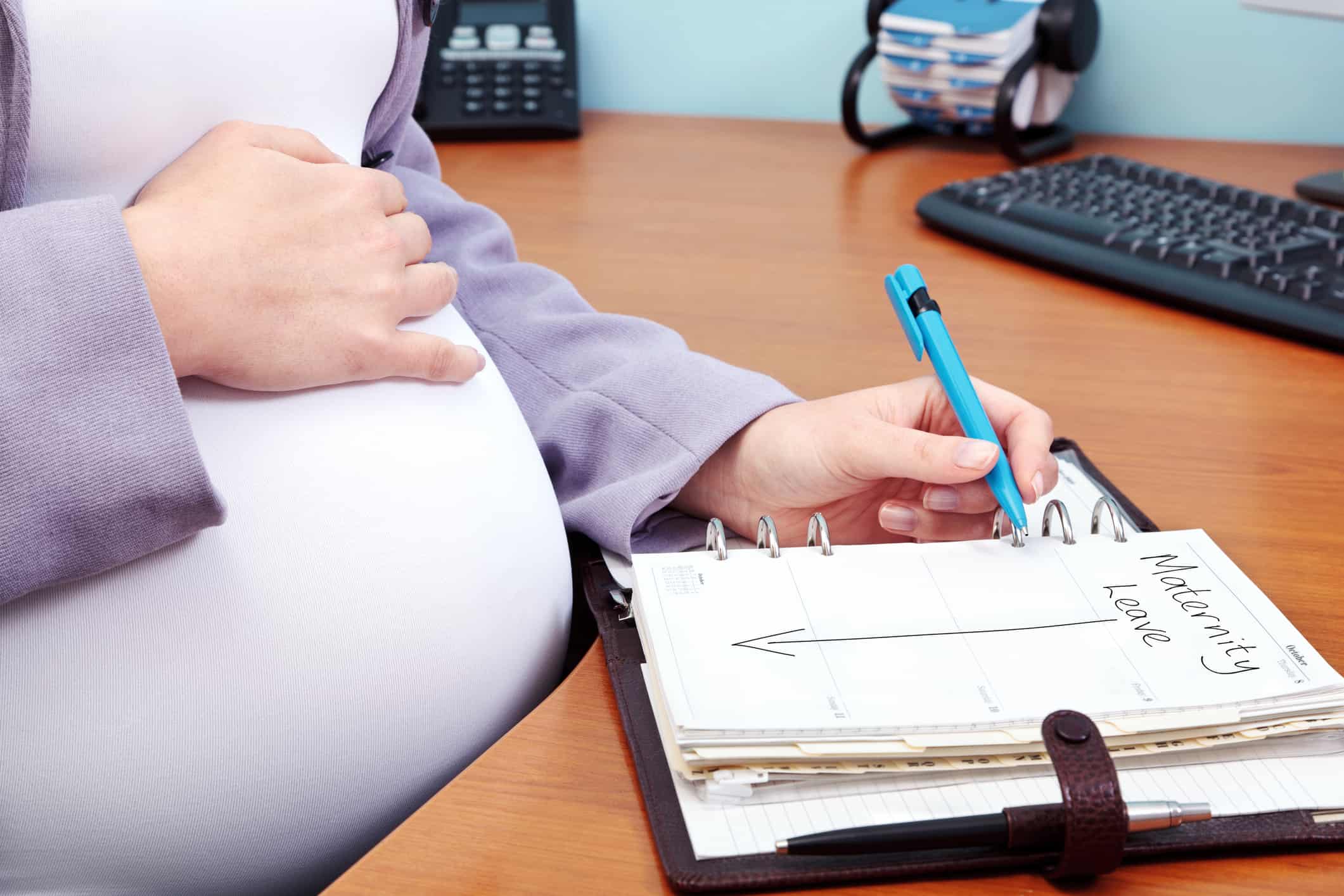 Photo of a pregnant woman in an office writing the words Maternity Leave in her diary.