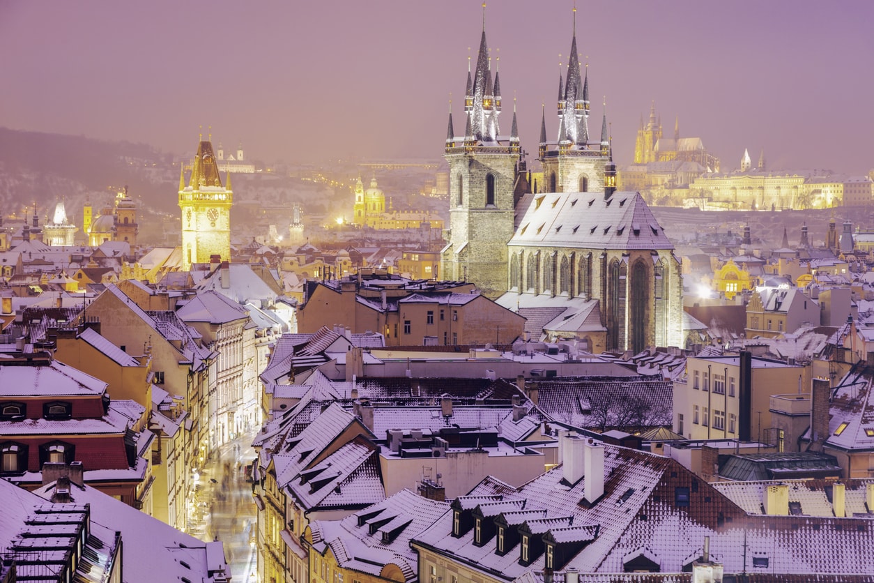 A snow-covered Prague winter cityscape with the Church of Our Lady Before Týn, Old Town Hall Clock Tower, and Prague Castle in the distant background.